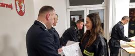 Na zdjęciu w sali konferencyjnej Komendy Powiatowej Państwowej Straży Pożarnej widoczny zastępca komendanta powiatowego Państwowej Straży Pożarnej w Gorlicach i Prezes Zarządu Oddziału Powiatowego Związku Ochotniczych Straży Pożarnych Rzeczypospolitej Polskiej w Gorlicach ubrani w ubrania galowe koloru granatowego wręczający zaświadczenia ukończenia szkolenia podstawowego strażaków ratowników OSP druhnie ubranej w ubranie koszarowe koloru czarnego. 