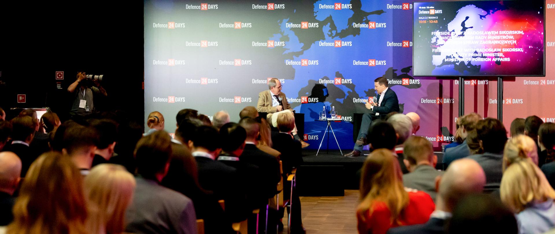 The photo shows two men sitting on stage. In front of them are a dozen or so people sitting in chairs. They are facing the stage. There is a map in the background. 
