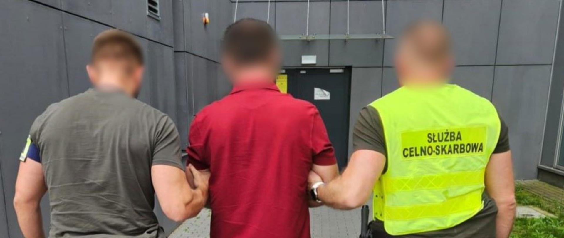 Trzech mężczyzn stojących tyłem, jeden ma kamizelkę z napisem służba celno-skarbowa. 