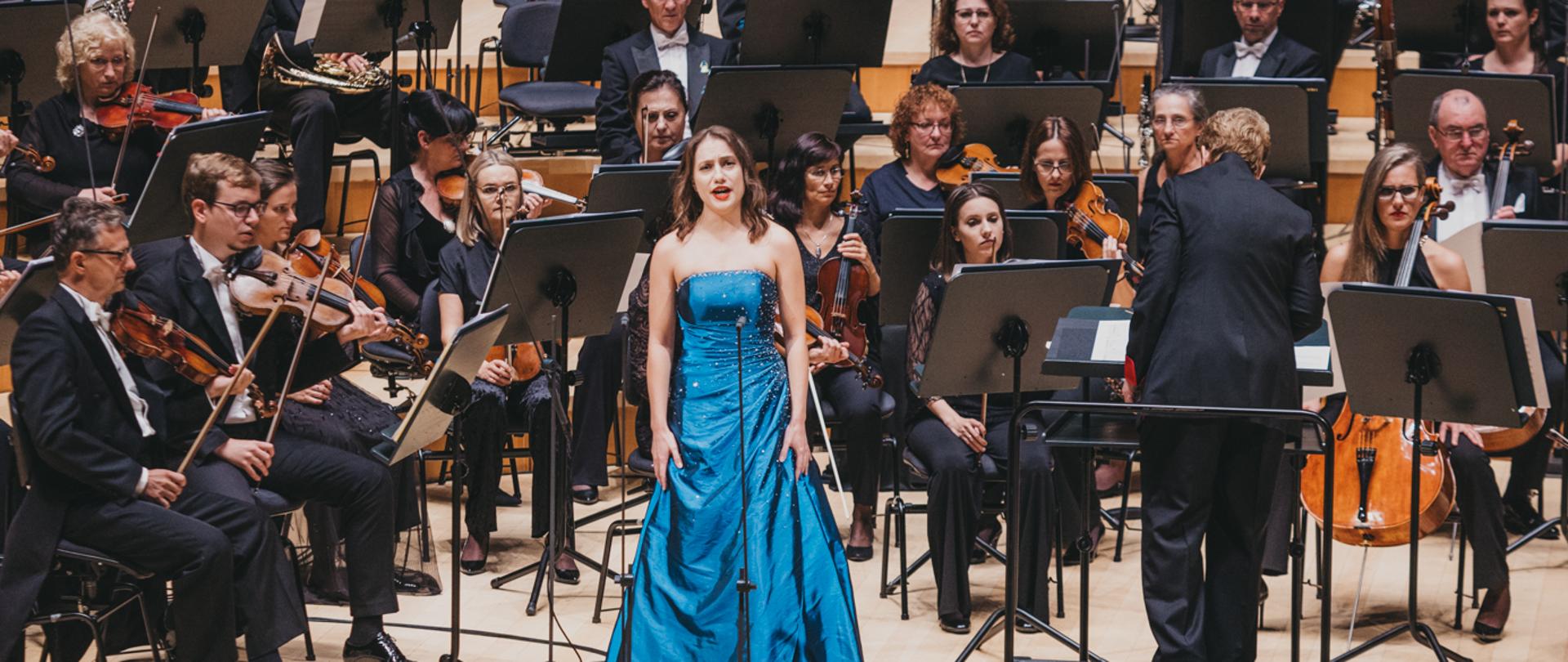 Kobieta śpiewa na estradzie, stojąc przed orkiestrą.