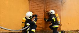 strażacy ubrani w sprzęt ODO z nawodniona linią strażacką otwierają pomieszczenie koloru żółtego z którego wydobywa się dym