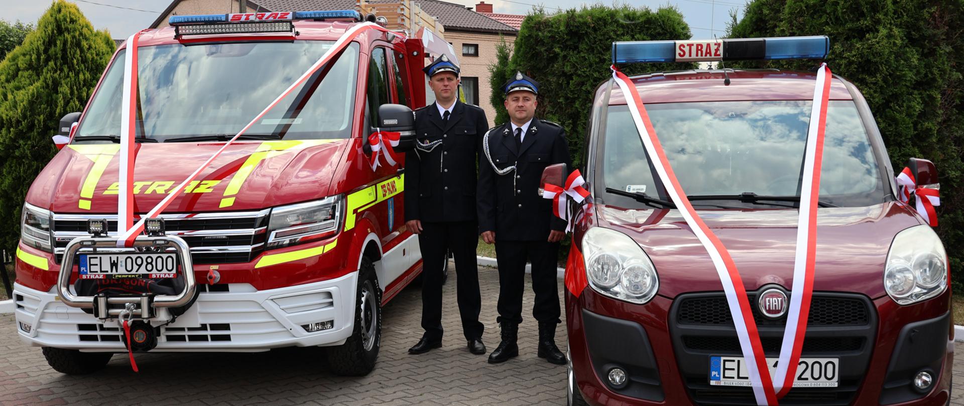 dwa samochody pożarnicze, po lewej lekki samochód ratowniczo-gaśniczy po prawej samochód operacyjny, pomiędzy nimi stoi dwóch druhów, w tle tuje i dom