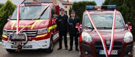 dwa samochody pożarnicze, po lewej lekki samochód ratowniczo-gaśniczy po prawej samochód operacyjny, pomiędzy nimi stoi dwóch druhów, w tle tuje i dom