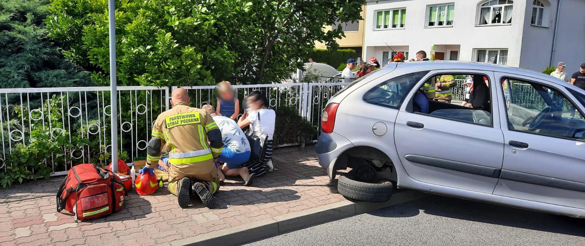 Fotografia przedstawia ujęcie chodnika przy ulicy Słowackiego. Na chodniku za jezdnię tyłem do fotografującego klęczy strażak w ubraniu specjalnym, ze zdjętym hełmem, naprzeciwko strażaka na chodniku siedzi i klęka kilka osób, w tym dzieci w wieku przedszkolnym. O prawej stronie kadru widoczny prawy bok samochodu citroen koloru srebrnego z urwanym tylnym kołem. W tle za chodnikiem drzewa i krzewy na prywatnej posesji. Jest dzień, słonecznie.