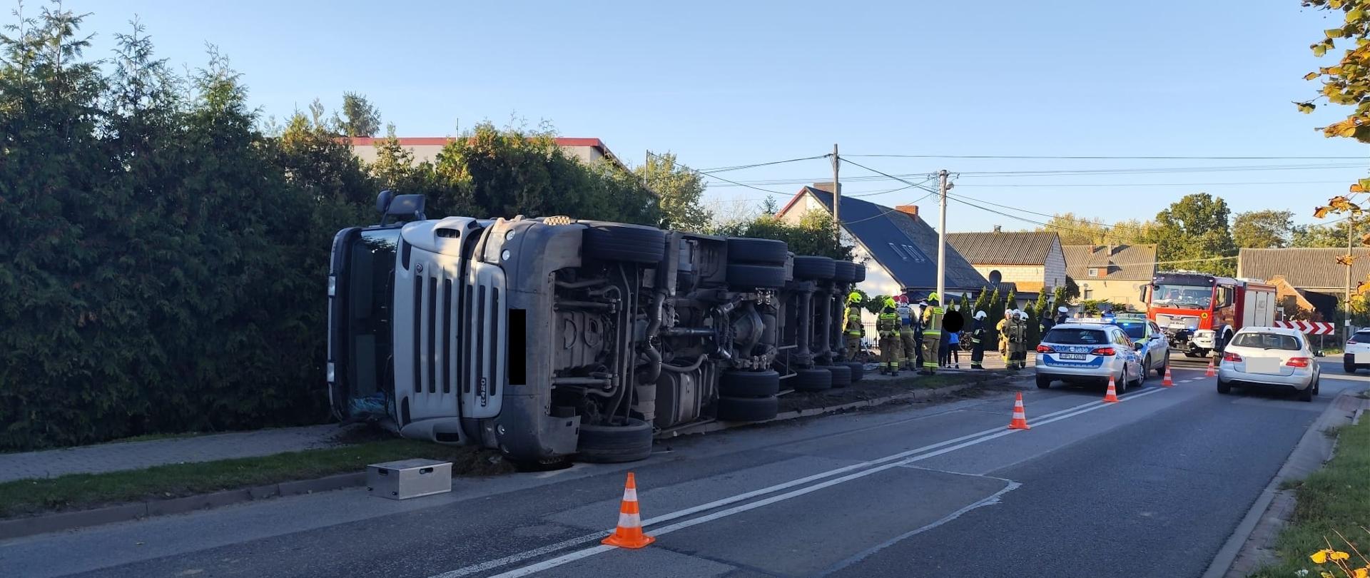 Pojazd ciężarowy leży na chodniku, w pobliżu strażacy i policja