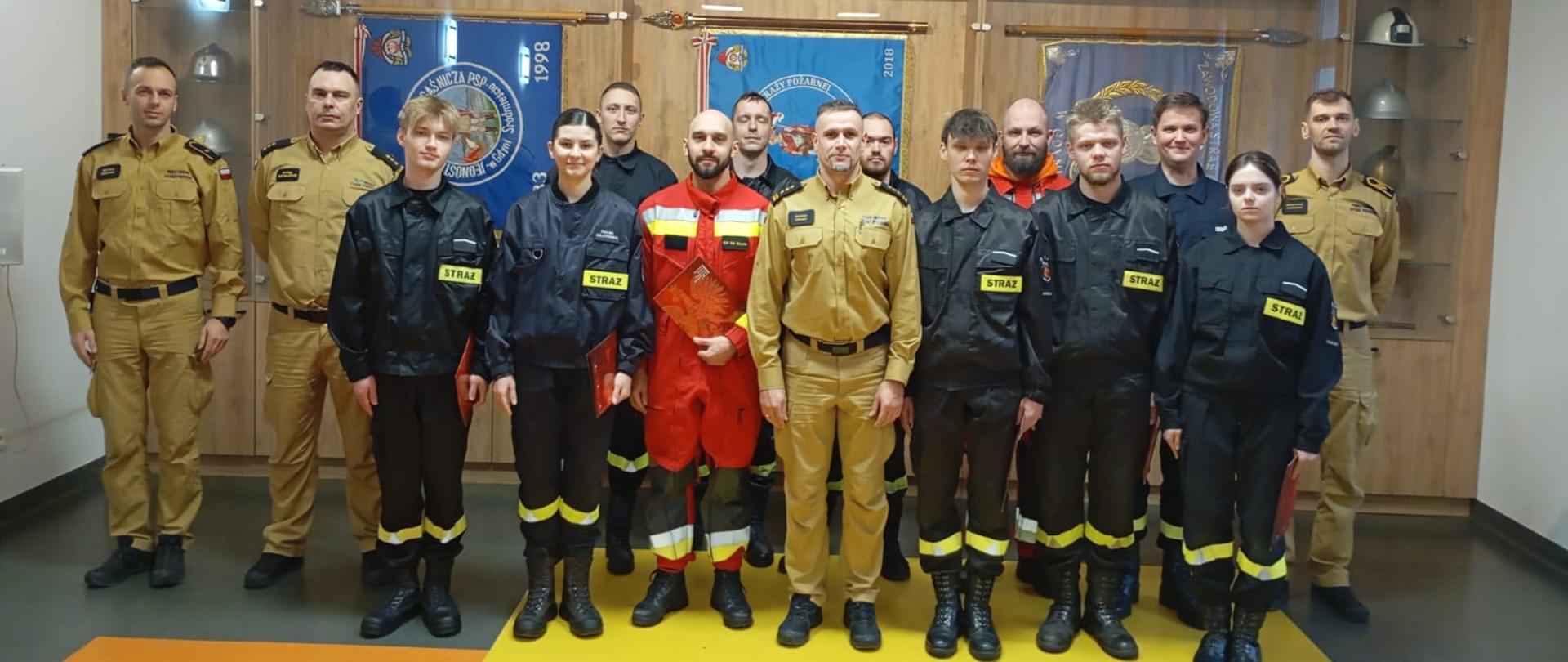 Na tle sztandarów stoi komendant, ze strażakami oraz adeptami kursu podstawowego strażaków ratowników ochotniczych straży pożarnych.