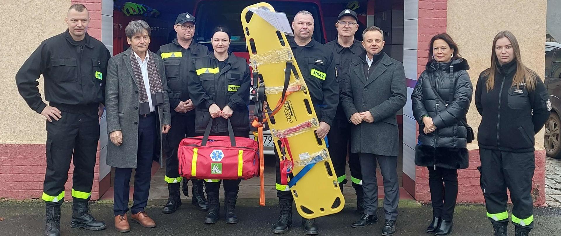 Zdjęcie grupowe przed budynkiem remizy na tle otwartej bramy garażowej z napisem OSP Mierczyce ponad nią. Przedstawiciele KRUS, Urzędu Gminy oraz strażacy prezentują wspólnie otrzymany czerwony plecak medyczny oraz żółtą ratowniczą deskę ortopedyczną.