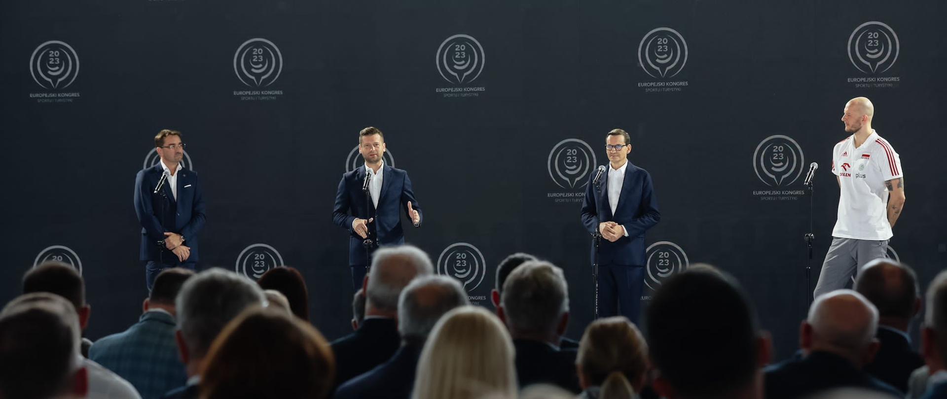 Four men standing on stage, the audience is listening. Prime Minister Mateusz Morawiecki together with Minister of Sport and Tourism Kamil Bortniczuk, President of the Polish Volleyball Federation Sebastian Świderski and captain of the Polish national volleyball team Bartosz Kurek announce that Poland will host the 2027 Volleyball World Championships