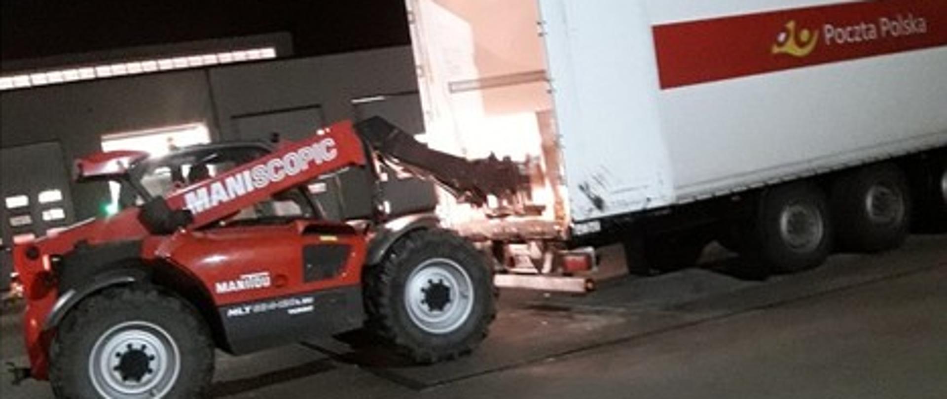 Zdjęcie przedstawia czerwoną ładowarkę marki MANITOU, podczas wyładowywania palety ze środkami ochrony indywidualnej z ciężarówki z logiem Poczta Polska.