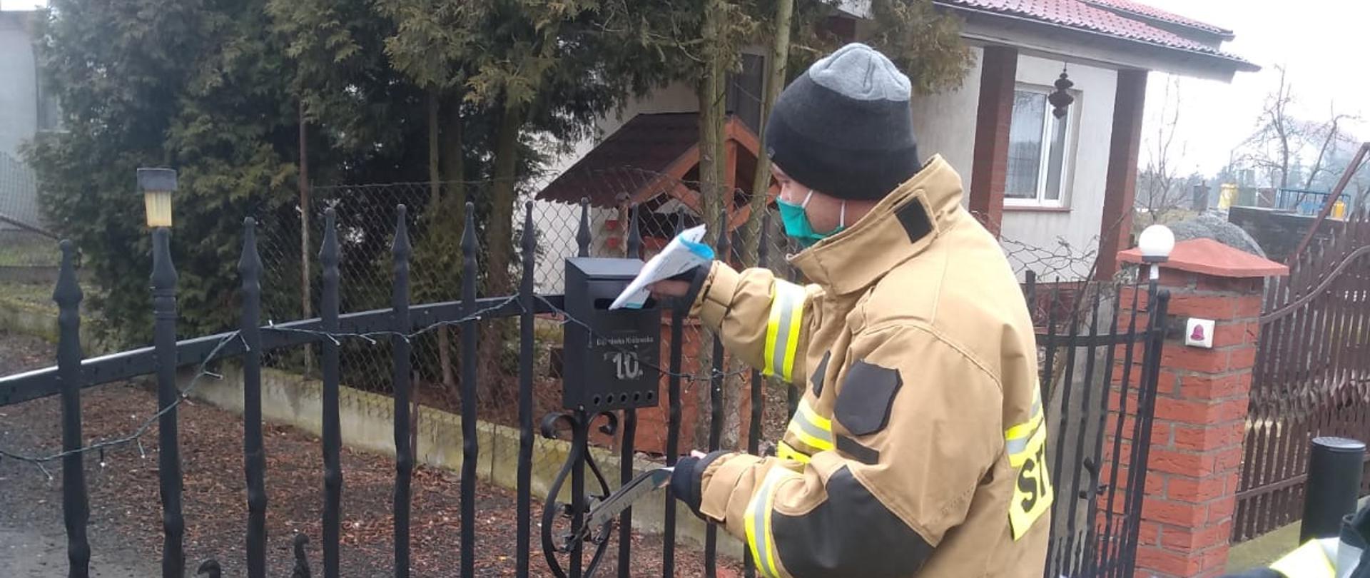 Zdjęcie przedstawia strażaka w piaskowym mundurze bojowym, który wkłada ulotki do skrzynki na listy zamontowanej na kutym ogrodzeniu. Strażak ma ubraną zieloną maseczkę. W tle drzewa i zabudowania.