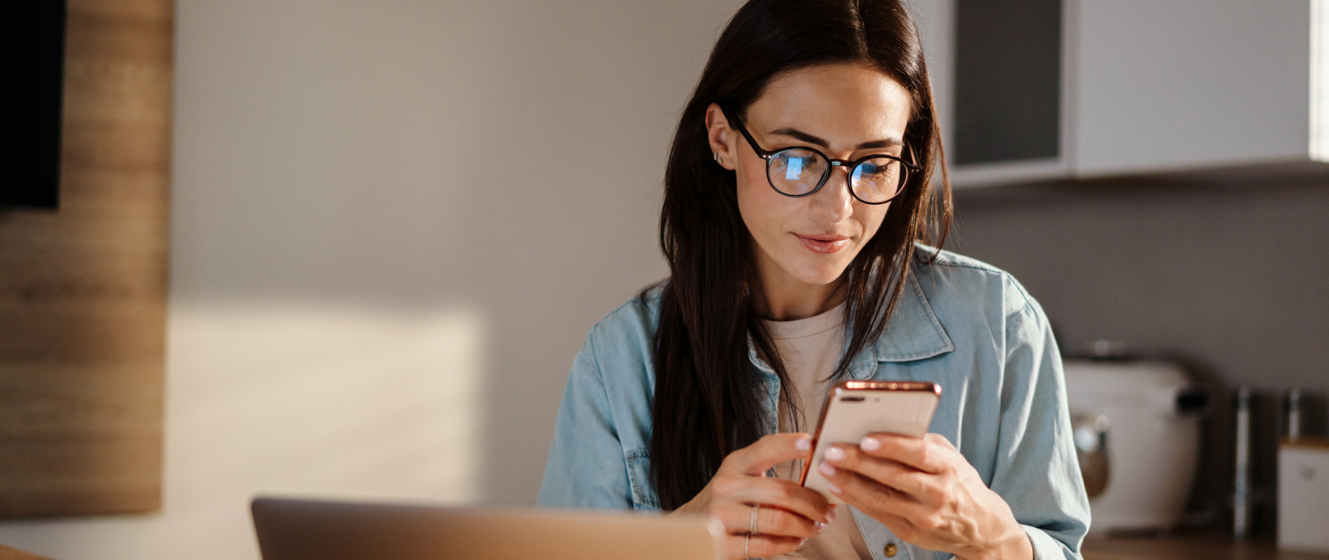 Kobieta w okularach siedzi w kuchni. W rękach trzyma telefon, na blacie stoi laptop.