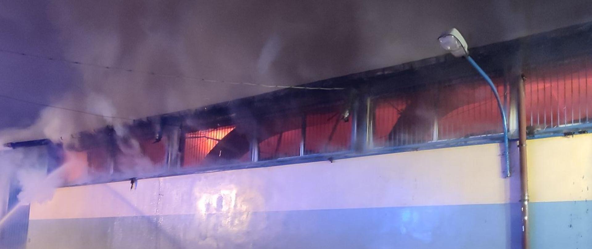 Ściana budynku, przez okna umieszczone w górnej części ściany widać ogień w środku budynku. Nad budynkiem gęsty dym. Na ziemi przed budynkiem leżą węże strażackie