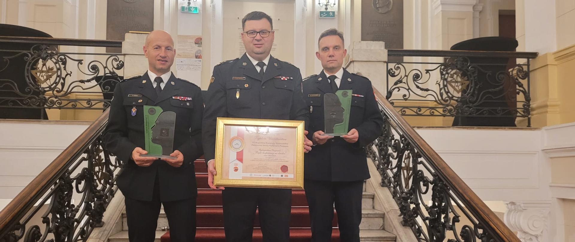 trzech strażaków stoi na schodach, dwóch z nich trzyma statuetki, mężczyzna w środku trzyyma duży dyplom