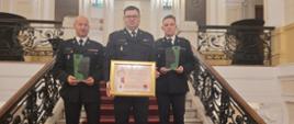 trzech strażaków stoi na schodach, dwóch z nich trzyma statuetki, mężczyzna w środku trzyyma duży dyplom