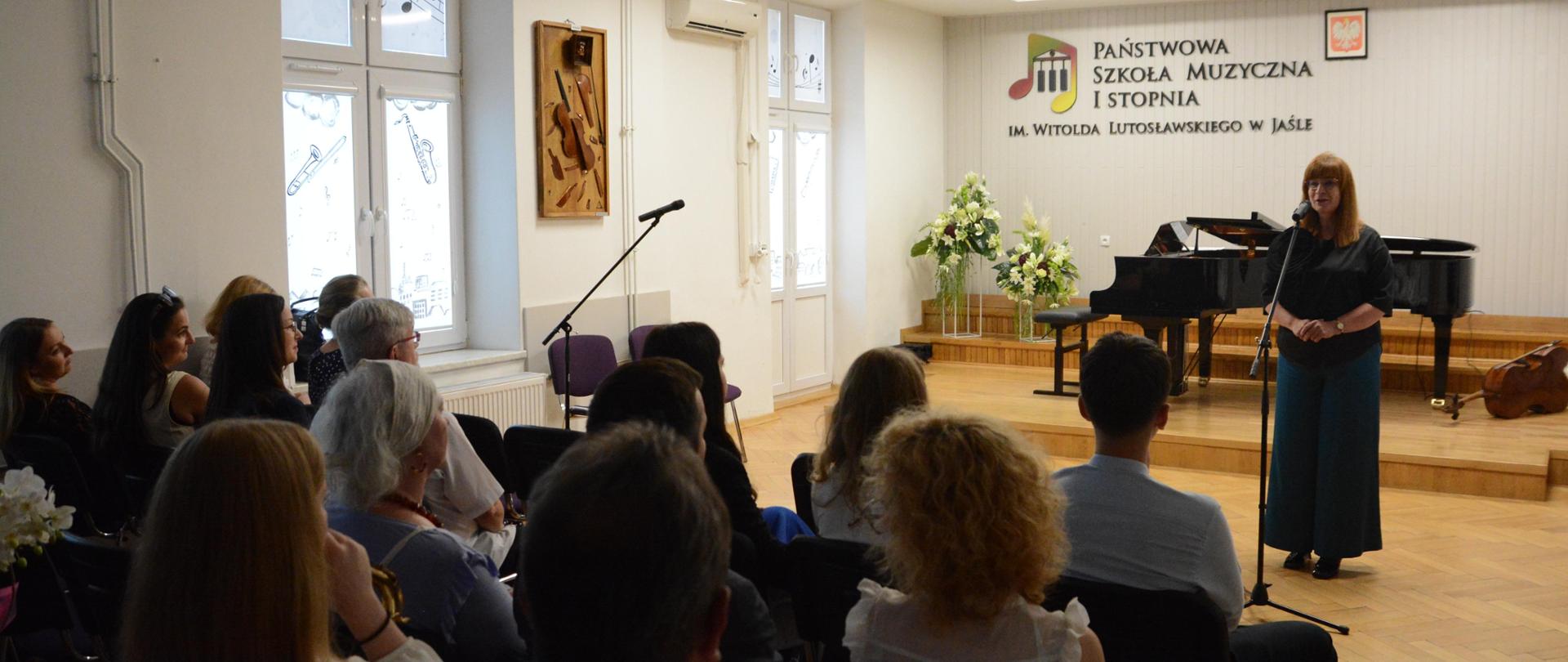 w auli PSM, podczas pożegnania klas kończących PSM I st. w Jaśle widnieje dyrektor szkoły, która przemawia do zgromadzonej społeczności szkolnej. Kolorystyka zdjęcia jest biało-żółto-szaro- brązowo-czarna.