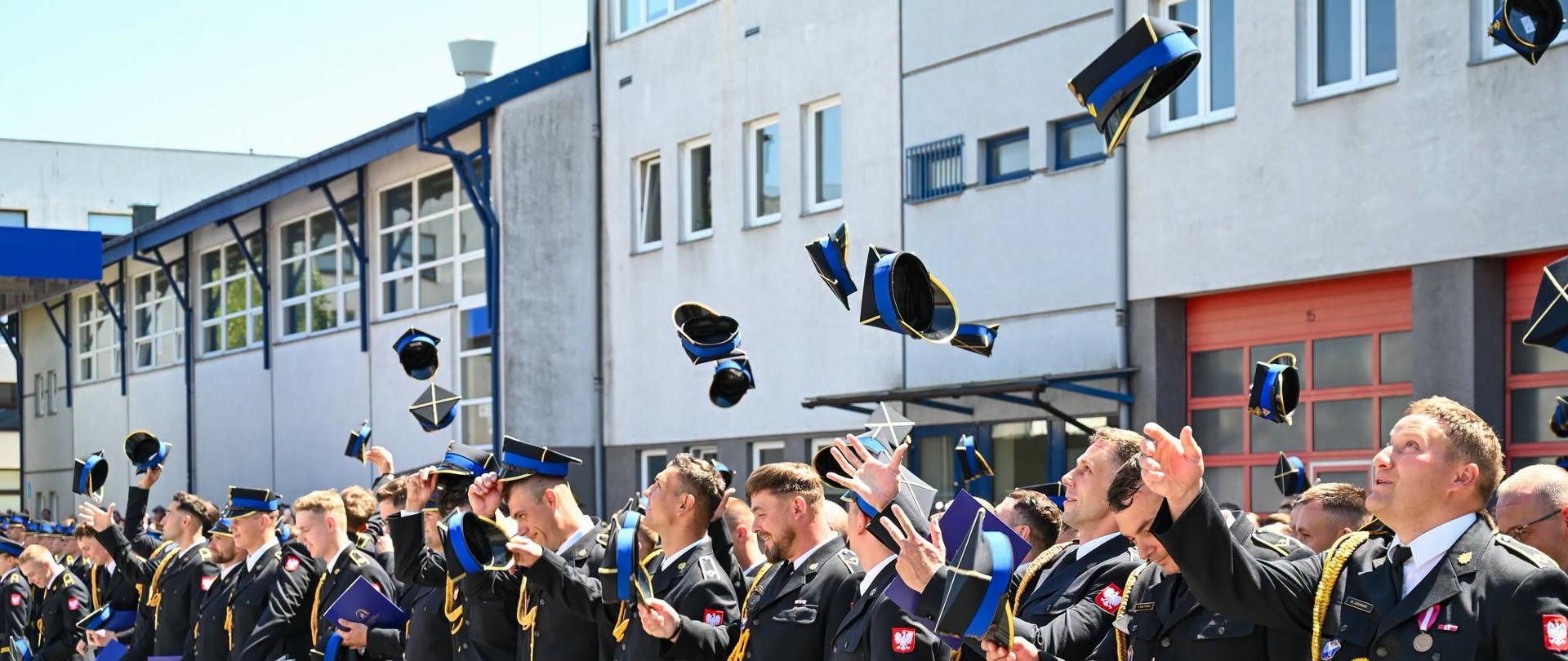 Grupa absolwentów w mundurach Państwowej Straży Pożarnej stoi w szeregu na placu przed budynkiem jednostki. Strażacy podrzucają w górę czapki rogatywki i trzymają w rękach niebieskie teczki z dokumentami. Zdjęcie przedstawia radosny moment zakończenia uroczystości promocyjnej.
