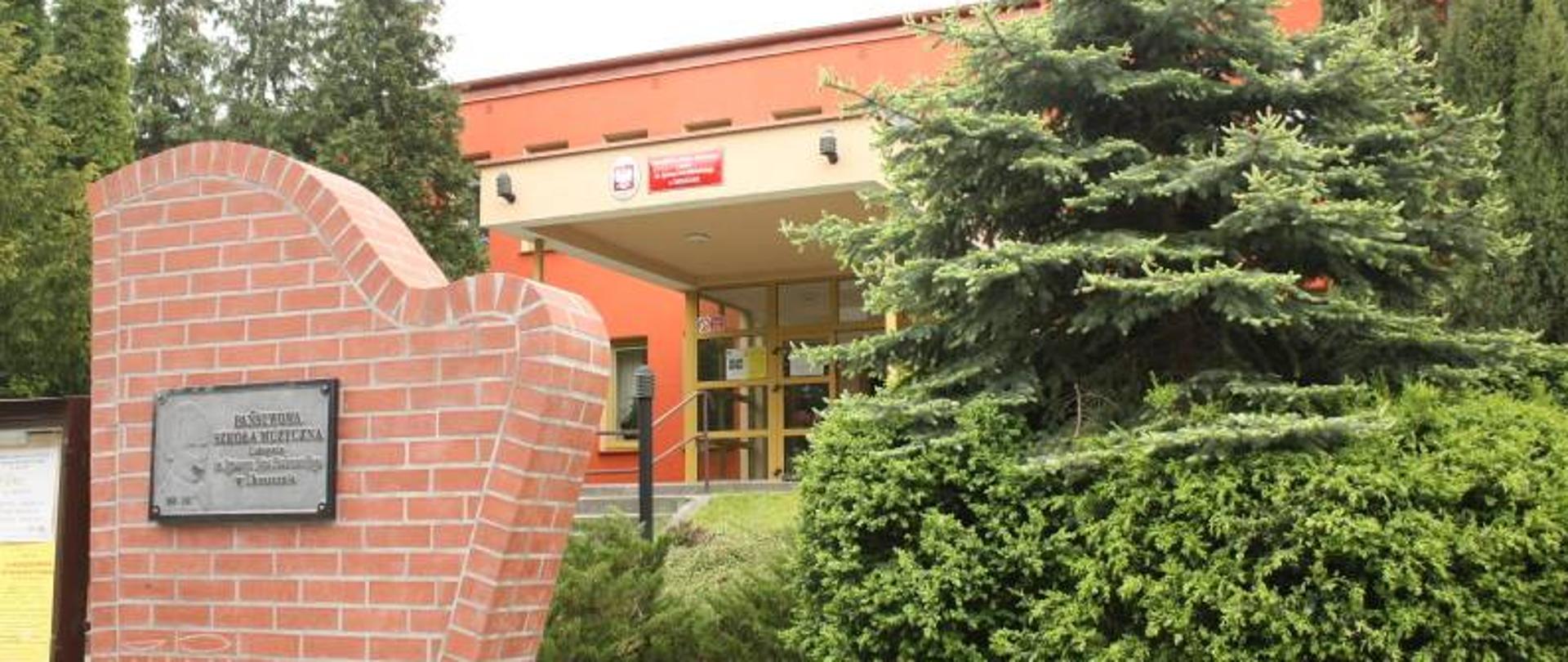 Fotografia, na niej widok na żółte drzwi wejściowe blado czerwonego koloru budynku szkoły muzycznej, przed brązowym metalowym płotem stoi murowany kształt harfy z zawieszoną na niej prostokątna tablica z nazwą szkoły z imieniem patrona na czarno szarym tle. Przed szkołą widać rosnące zielone drzewa i krzewy. Fotografia z 2019 r.