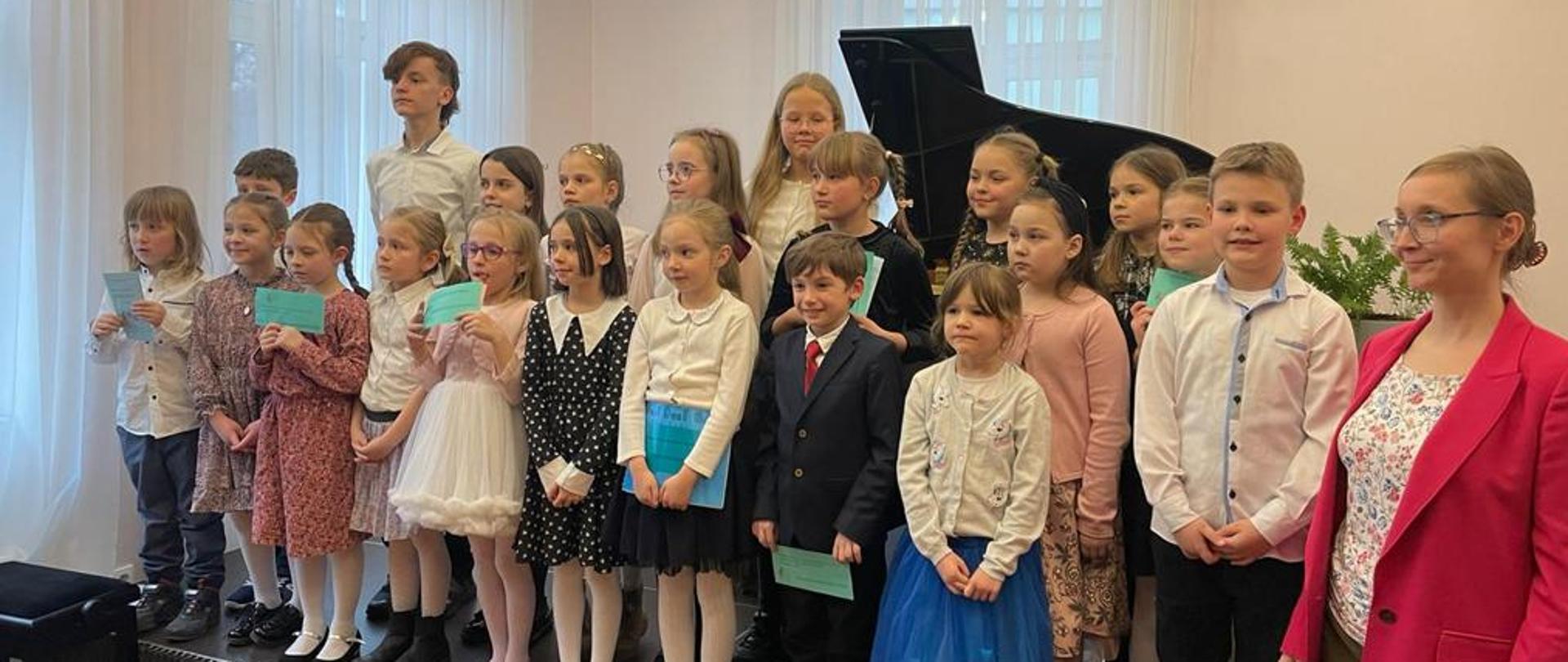 Grupowe zdjęcie dzieci, wraz z nauczycielem Panią A. Strzygocką, dzieci stoją na scenie w auli PSM I i II st. w Bystrzycy Kłodzkiej