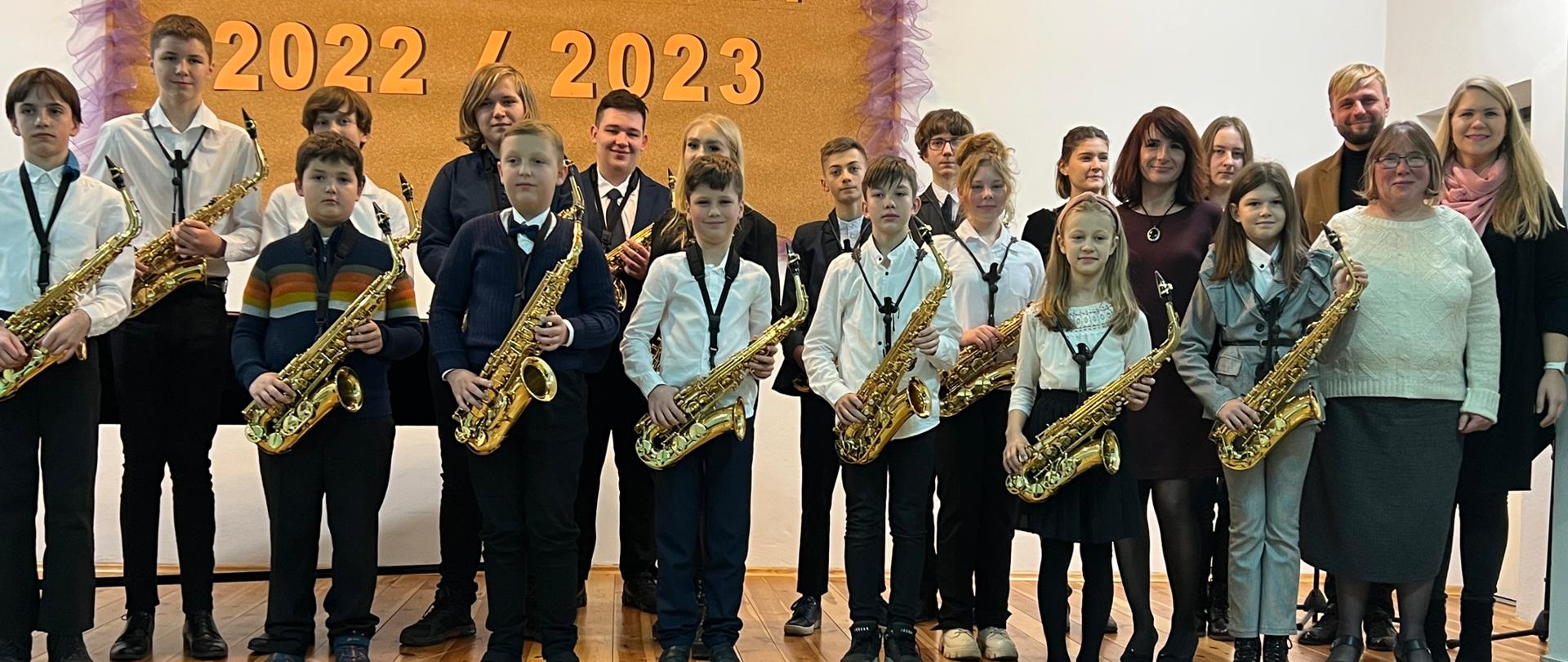Zdjęcie przedstawiające wszystkich wykonawców koncertu muzyki saksofonowej na scenie w auli szkoły.