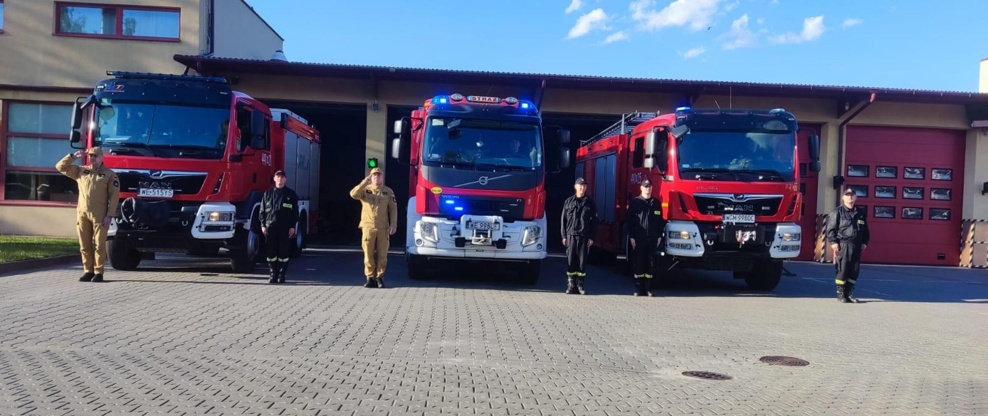 Strażacy oddają hołd dla śp. druha Marcina Raczyka z OSP Żerniki (powiat kaliski, woj. wielkopolskie). Strażacy stoja przed garażem JRG za nimi stoja samochody pożarnicze z włączonymi sygnałami świetlnymi i dźwiękowymi