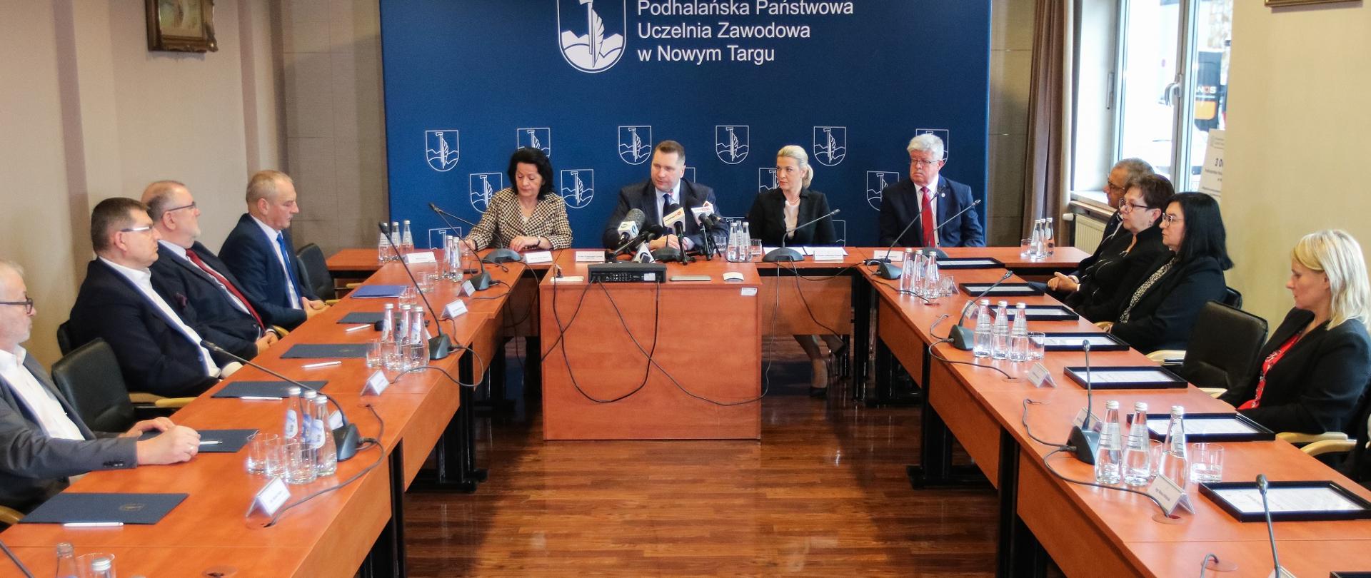 Widok na salę przy ustawionych w czworokąt stołach siedzą elegancko ubrani ludzie, u szczytu stołu minister Czarnek, za nim ścianka z napisem Podhalańska Państwowa Uczelnia Zawodowa w Nowym Targu, po lewej okna.