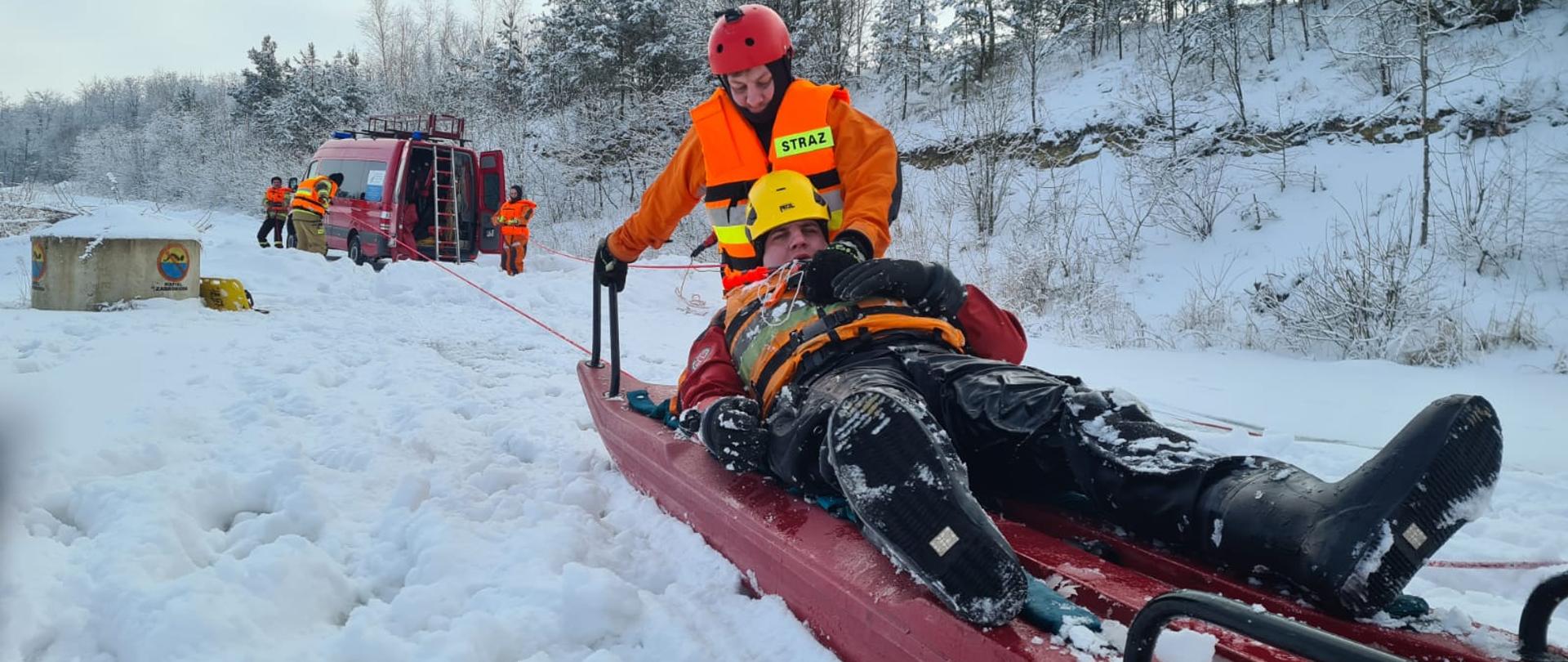 Zdjęcie przedstawia ratownika ubranego w ubranie specjalne do działań na akwenach wodnych. Ratownik wraz z osobą wydobytą z przerębli znajduje się na saniach służących do prowadzenia działań ratowniczych na lodzie.
W oddali widać ratowników i pojazd strażacki w kolorze czerwonym. Ratownicy za pomocą liny ciągną strażaka wraz z pozorantem w swoja stronę, w bezpieczne miejsce. Teren dookoła porywa śnieg.
