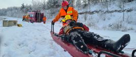 Zdjęcie przedstawia ratownika ubranego w ubranie specjalne do działań na akwenach wodnych. Ratownik wraz z osobą wydobytą z przerębli znajduje się na saniach służących do prowadzenia działań ratowniczych na lodzie.
W oddali widać ratowników i pojazd strażacki w kolorze czerwonym. Ratownicy za pomocą liny ciągną strażaka wraz z pozorantem w swoja stronę, w bezpieczne miejsce. Teren dookoła porywa śnieg.
