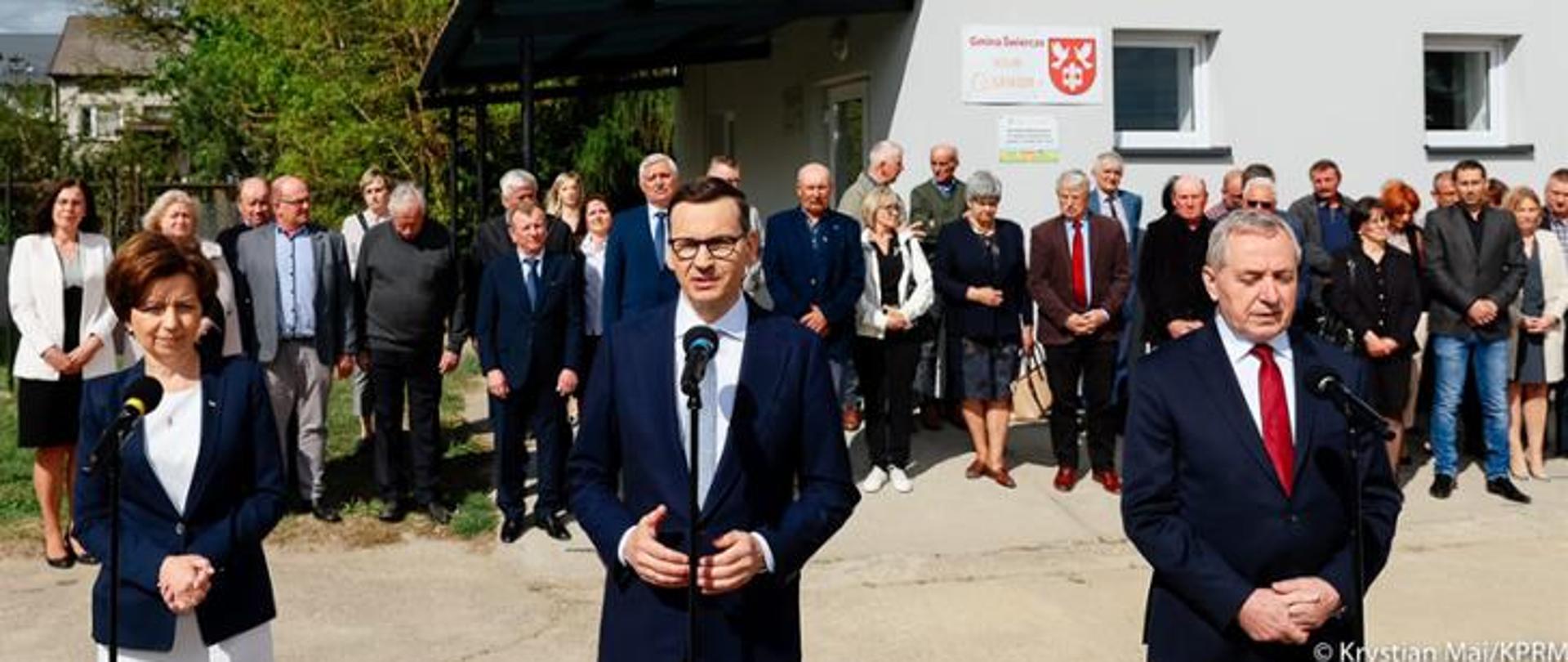 Dwóch mężczyzn i kobieta stojący przed mikrofonami. Za nimi wiele osób