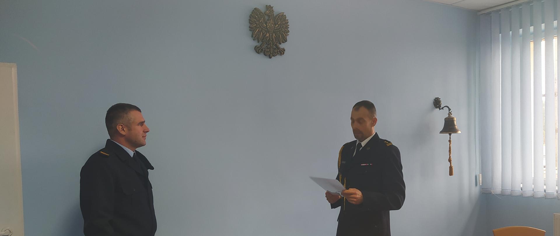 Zdjęcie wykonano w pomieszceniu w którym kolor ścian jest miebieski
przedstawia ono dwóch funkcjonariuszy
mężczyzna po prawej stronie jest w umundurowaniu wyjściowym z sznurem w stopniu brygadiera
drugi z mężczyzn jest w umundurowaniu służbowym w stopniu sekcyjnego.
Mężczyzna z wyższym stopniem trzyma kartkę papieru. Nad ich głowani
wisi godło.