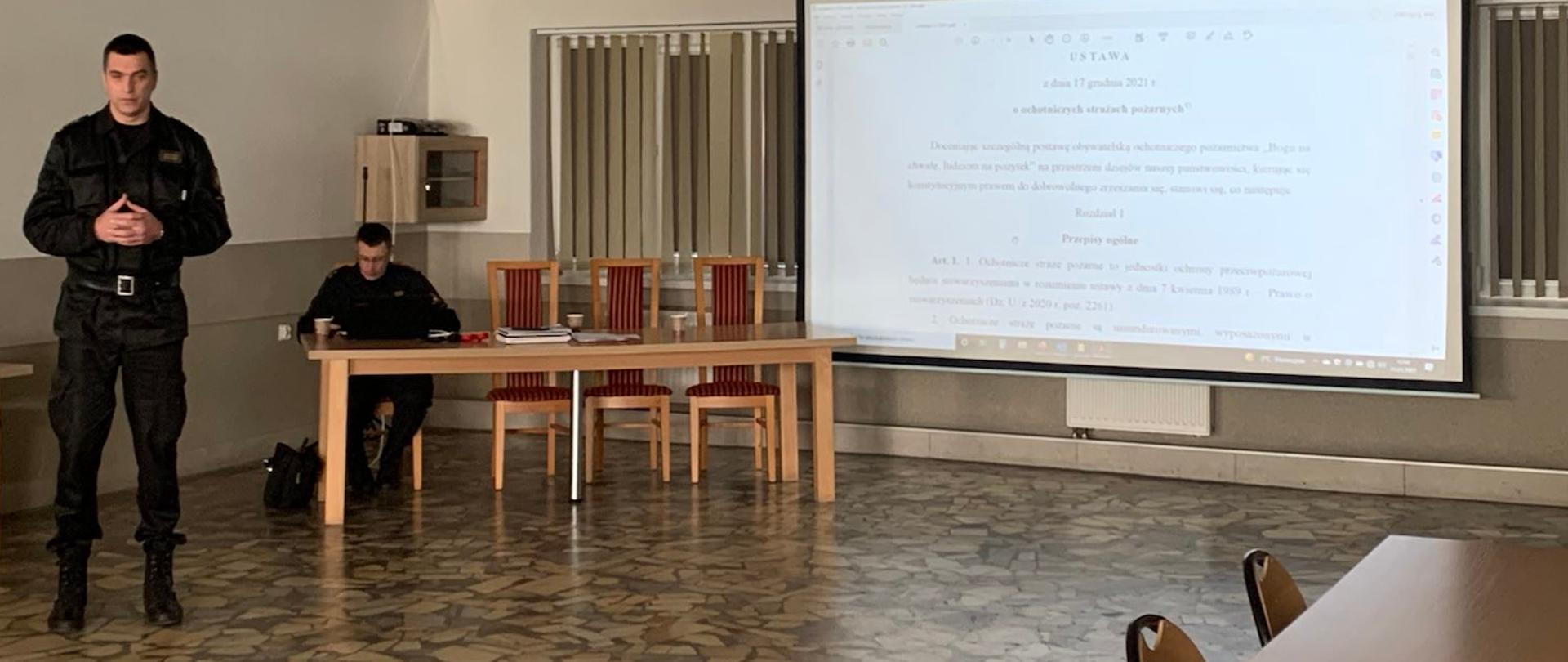 Na zdjęciu widać Komendanta Powiatowego PSP w Oleśnicy w pozycji stojącej, który prowadzi spotkanie w sprawie Ustawy o OSP. Na ekranie rzutnik wyświetla treść ustawy. 