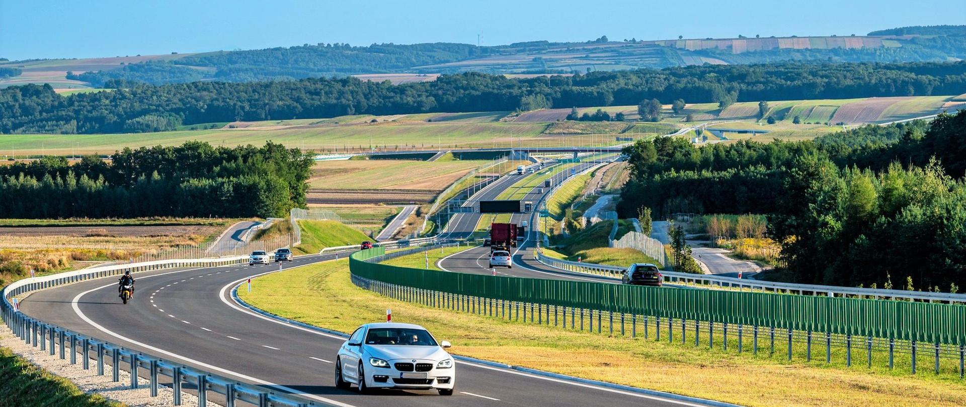Droga ekspresowa S7 na odcinku Moczydło - Miechów. Dwujezdniowa droga ekspresowa wijąca się między polami i lasami po niewielkich pagórkach. Widoczne pojazdy poruszające się w obu kierunkach.
