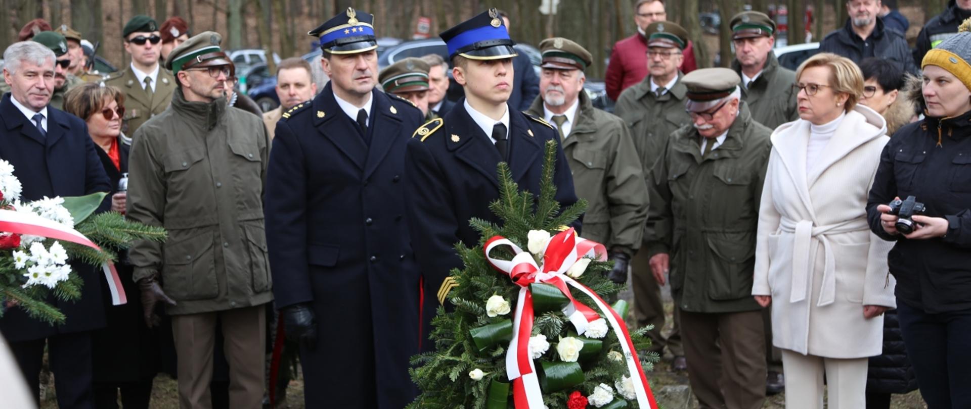 Na pierwszym planie kadet Centralnej Szkoły Państwowej Straży Pożarnej w Częstochowie trzymający wieniec kwiatów, za nim stojący Komendant Centralnej Szkoły PSP. Dookoła goście uroczystości