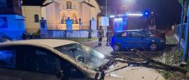 Na zdjęciu w porze nocnej widać uszkodzony samochód osobowy marki Opel przez konary drzewa. W tle widać samochód gaśniczy, strażaków oraz kościół.