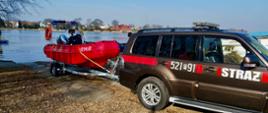 Samochód strażacki z napisem „STRAŻ” stoi nad brzegiem jeziora i ciągnie na przyczepie czerwoną łódź ratowniczą z silnikiem. W tle widać spokojną wodę, drzewa bez liści oraz zabudowania po drugiej stronie jeziora w słoneczny dzień. 
