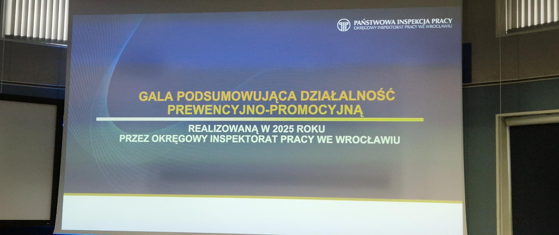 Slajd wyświetlony na ekranie podczas wydarzenia organizowanego przez Okręgowy Inspektorat Państwowej Inspekcji Pracy we Wrocławiu zawierający tytuł "Gala podsumowująca działalność prewencyjno - promocyjną realizowaną w 2025 roku przez Okręgowy Inspektorat Pracy we Wrocławiu"