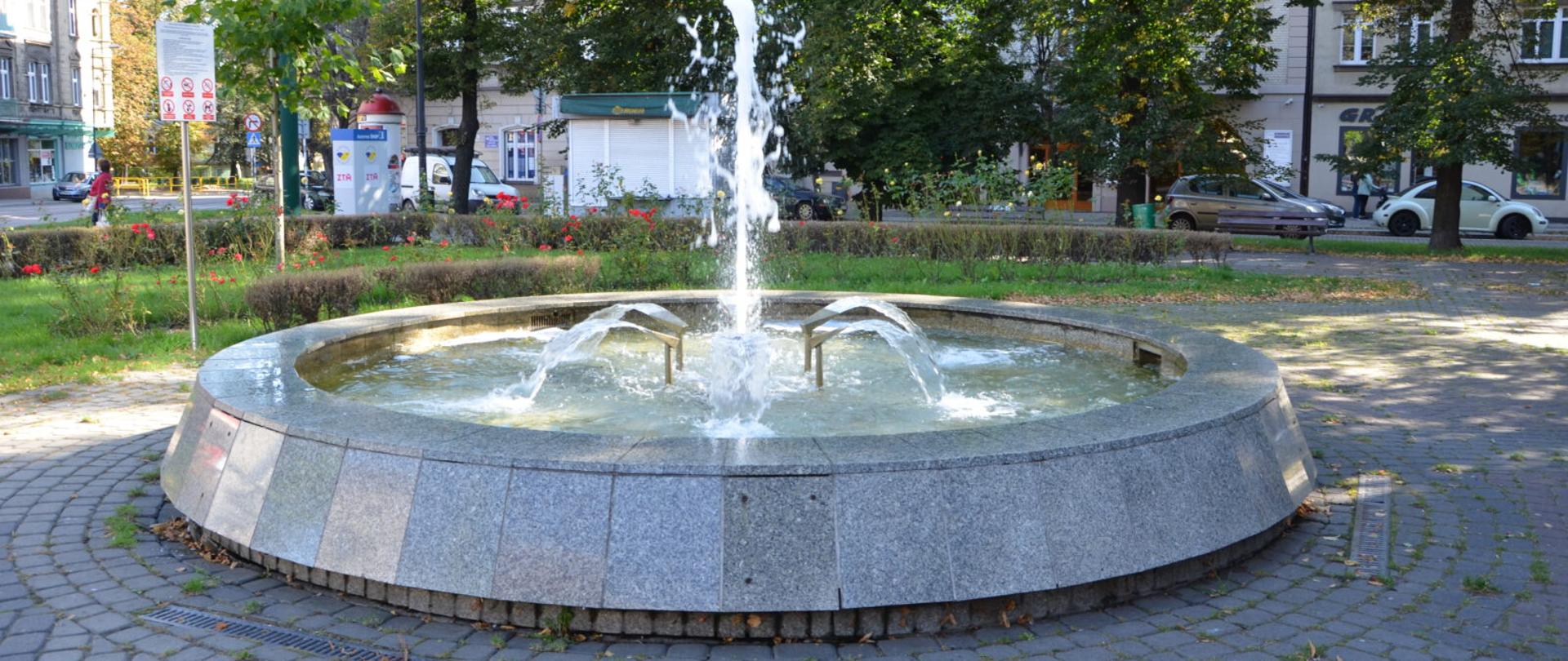 zdjęcie przedstawia fontannę na tle zieleń i bloki