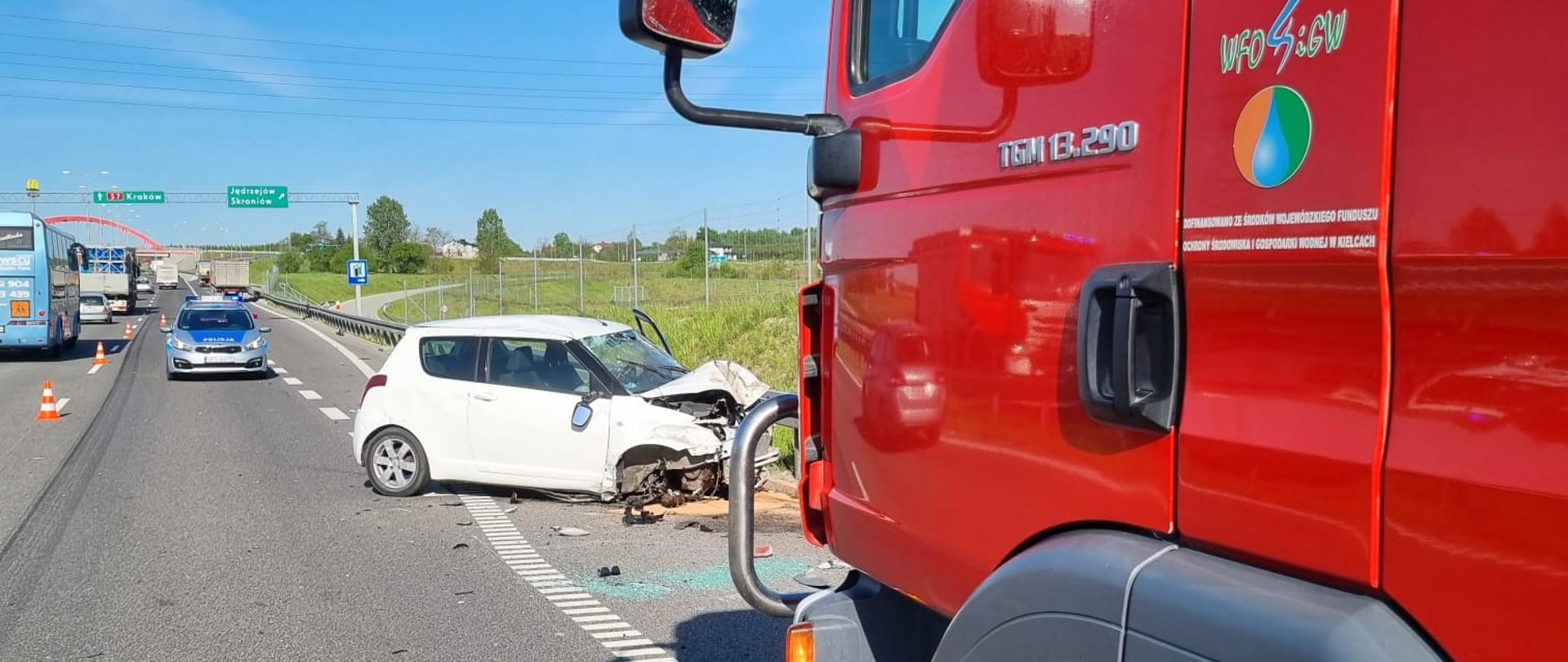Na zdjęciu widać po prawej stronie przód samochodu ratowniczo-gaśniczego. Pozostała część zdjęcia przedstawia drogę ekspresową, radiowóz policyjny oraz uszkodzony pojazd osobowy w białym kolorze, który brał udział w zdarzeniu. 