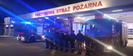 Zdjecie przedstawia strażaków stojących przed garażami i trzema pojazdami pożarniczymi z włączonymi białymi i niebieskimi światłami