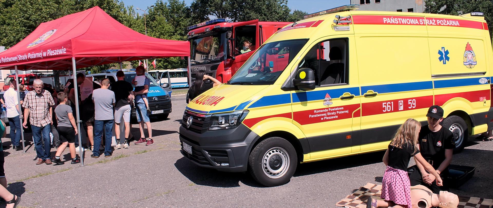 Grupa dzieci i dorosłych stojące przed stoiskiem edukacyjnym PSP, widoczny czerwony namiot stoiska z napisem Komenda Powiatowa Państwowej Straży Pożarnej w Pleszewie. Na pierwszym planie widoczna żólta karatka z napisem Państwowa Straż Pożarna, przed nią ćwicząca na manekinia kilkuletnia dziewczynka i strażak