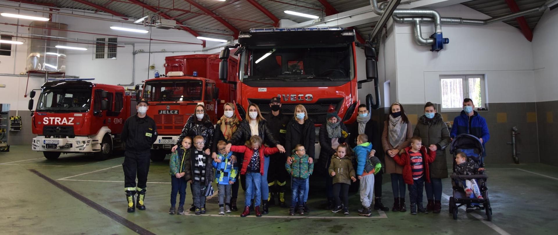 Grupa dzieci wraz z opiekunami i strażakami w ubraniach koszarowych stojących na tle wozu bojowego.