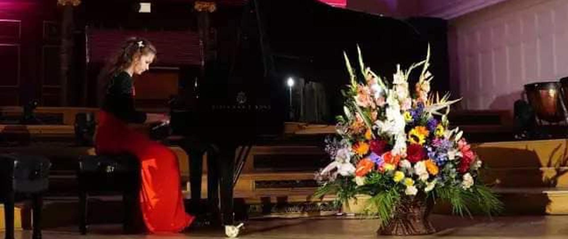 Pianistka przy fortepianie w czerwono-czarnej sukni. Przed fortepianem wielki kosz kwiatów, w tle prospekt organów.