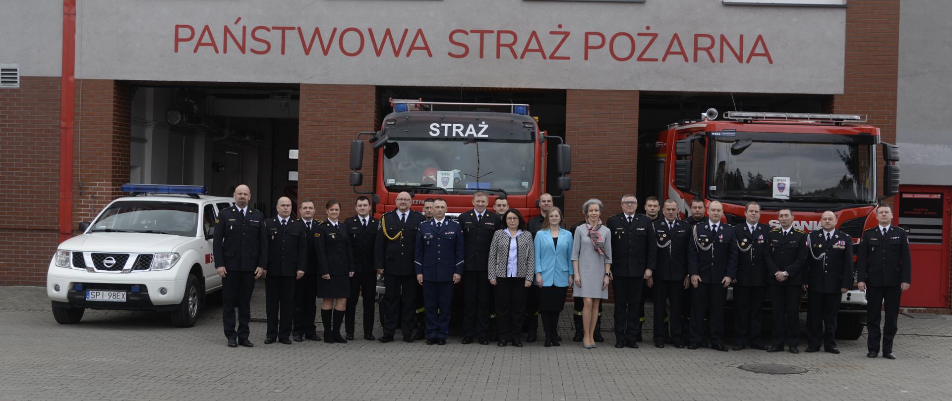 Uczestnicy odprawy pozują do zdjęcia na tle obiektu komendy miejskiej oraz samochodów ratowniczo - gaśniczych.