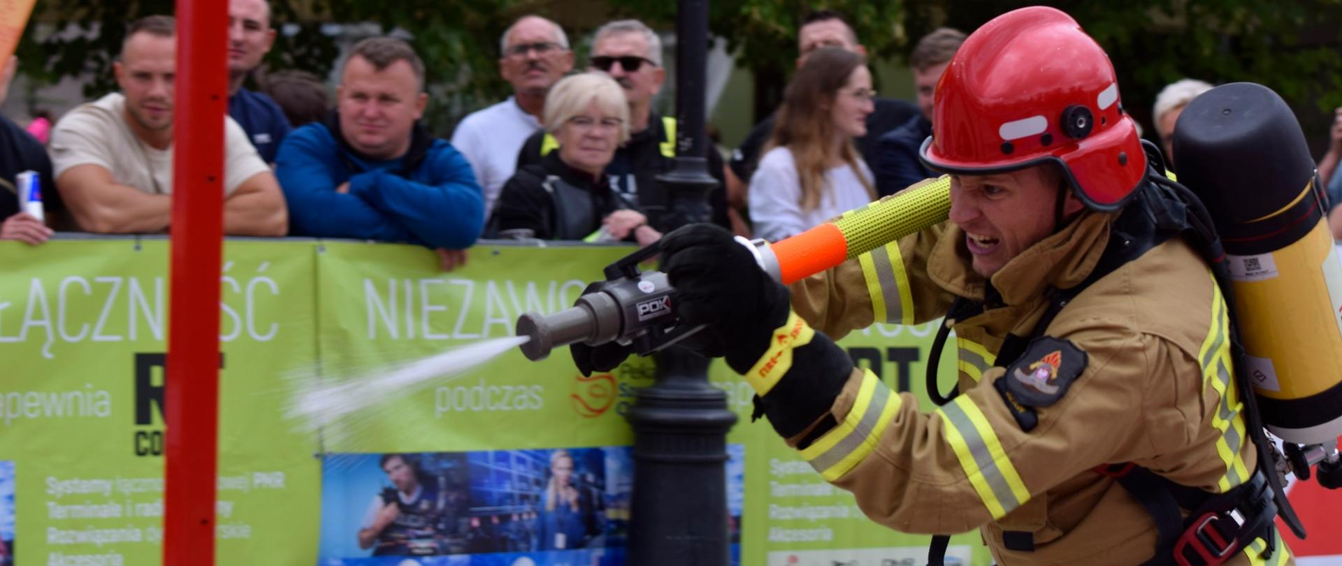 pora dzienna strażak w mundurze bojowym z wężem na plecach biegnie oddając strzał z prądownicy wodnej do celu, w tle widać kibicujących ludzi.