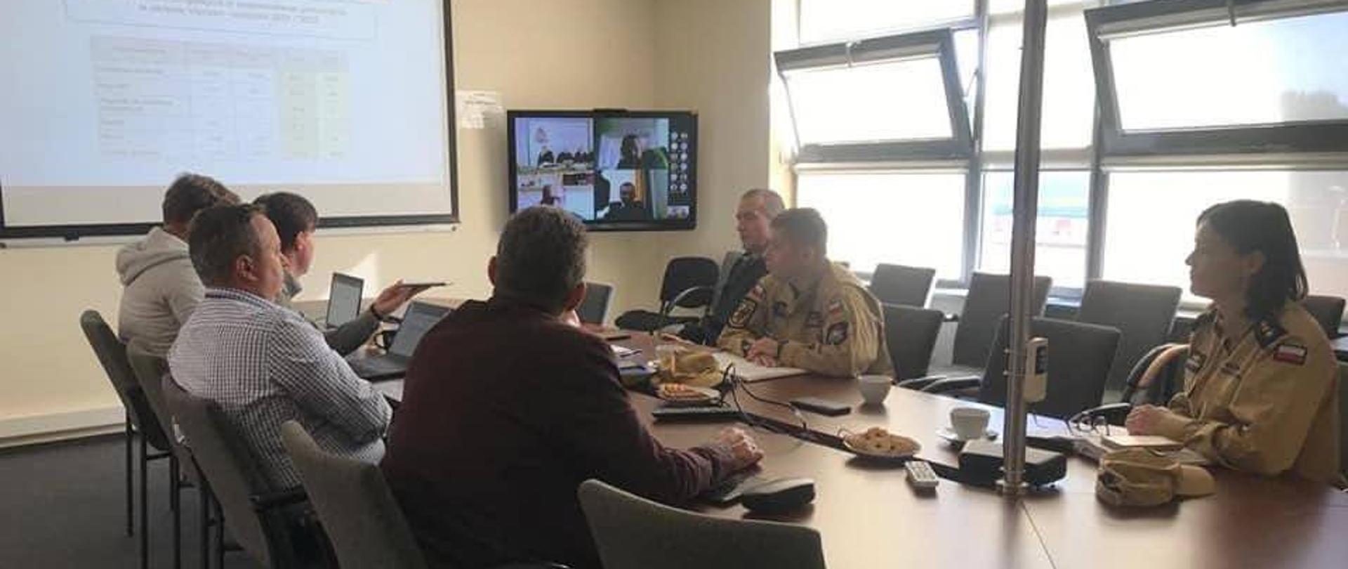 Przedstawiciele operatora autostrady oraz funkcjonariusze Państwowej Straży Pożarnej siedzą za stołem naprzeciwko siebie na ekranie wyświetlane są statystyki.