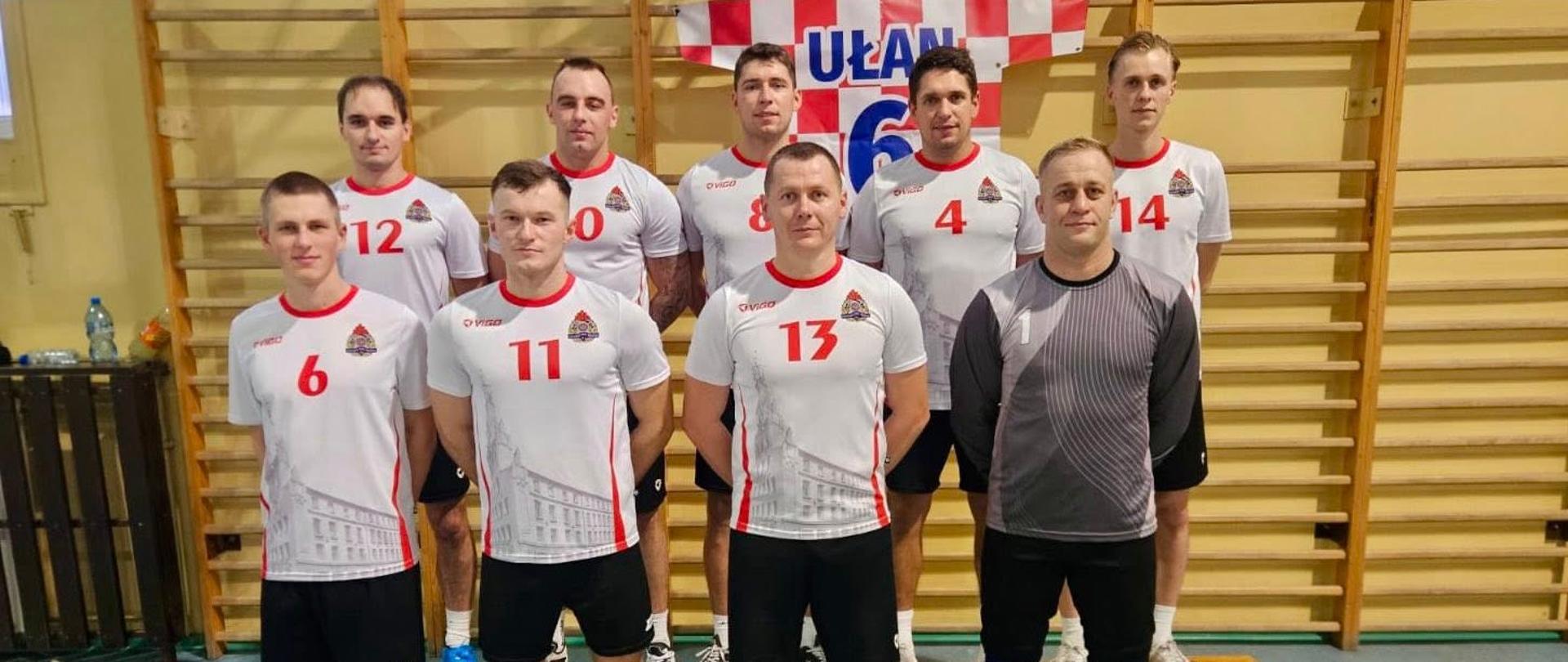 Zdjęcie przedstawia strażaków ustawionych całą drużyną w dwóch szeregach, na hali sportowej, w ubraniach sportowych, a przed nimi stoją puchary i dyplomy.