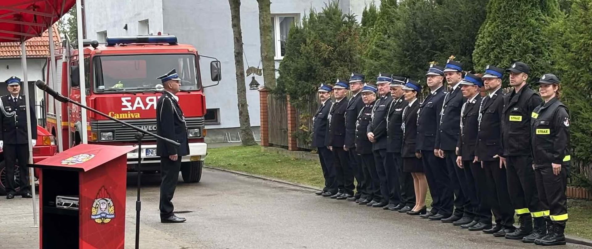 Strażak w mundurze wyjściowym stoi na przeciwko druhów ochotniczych straży pożarnych. W tle widoczny pojazd pożarniczy i czerwony, strażacki namiot. Dalej widoczne miejskie zabudowania.