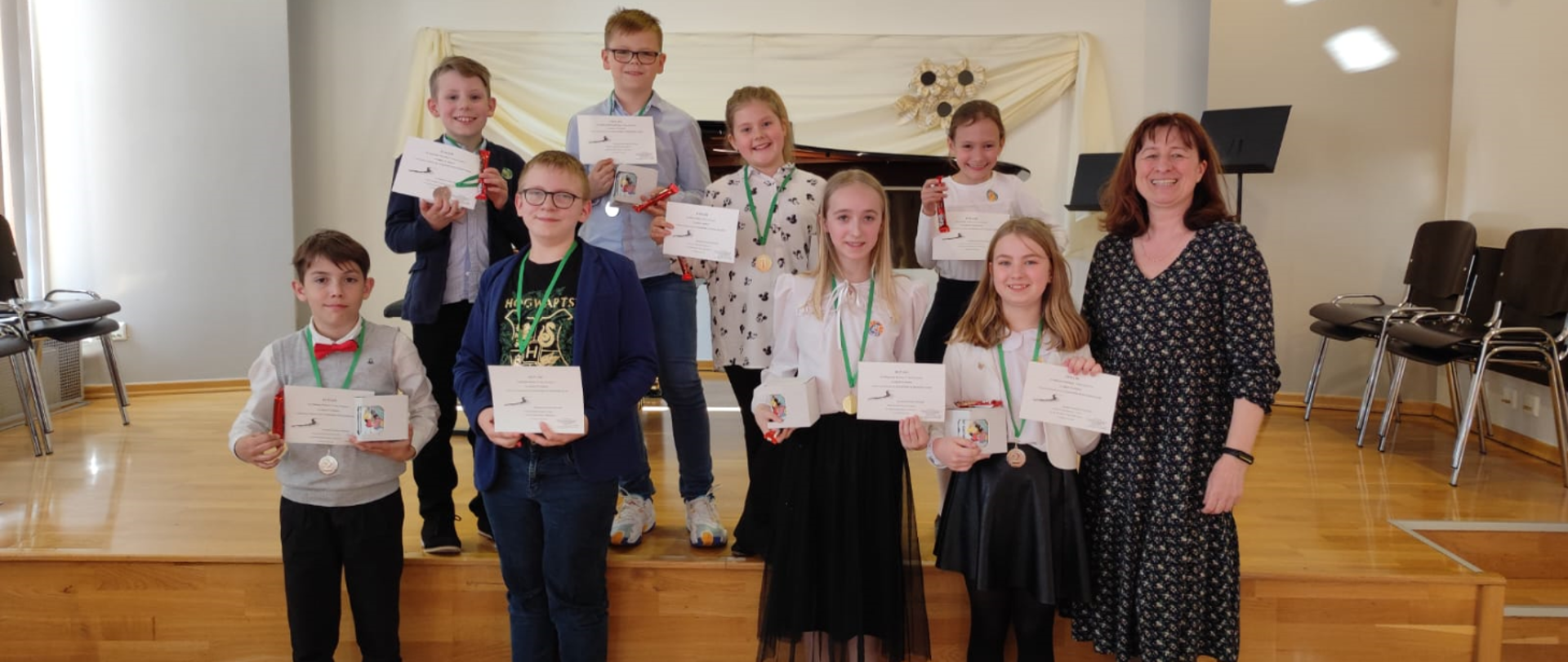 Na zdjęciu grupa uczniów wraz z nauczycielką- brunetką w ciemnej sukience. W tle otwarty fortepian. Wszyscy uczniowie trzymają dyplom oraz kubek w pudełku. na szyjach mają zawieszone medale.