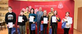 Laureaci turnieju na tle ścianki z koloru czerwonego w obecności dwóch strażaków oraz pana w garniturze