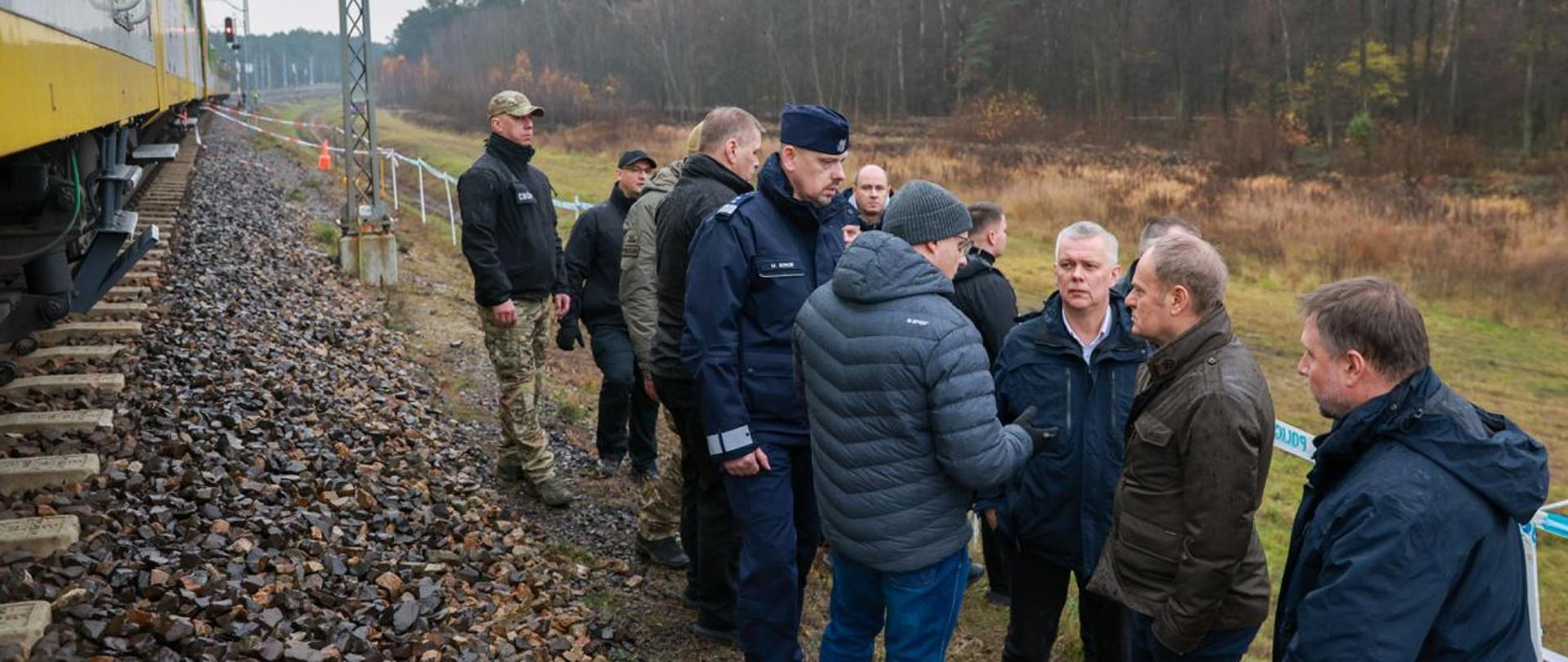 Po lewej stronie stoi na torach Kolej Mazowiecka, po prawej premier Donald Tusk, minister Tomasz Siemoniak, minister Marcin Kierwiński oraz przedstawiciele służb. W tle las.
