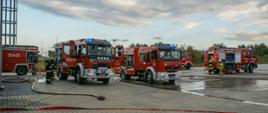 Pojazdy pożarnicze ustawione na placu zakładu po przeprowadzeniu ćwiczeń, w tle nieużytki rolne. Strażacy w ubraniach specjalnych stoją obok pojazdów.
